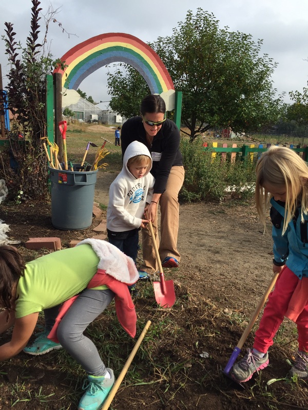 Students dig a garden bed with shovels and a parent volunteer, under the rainbow arch of the Columbine garden, with the greenhouse behind