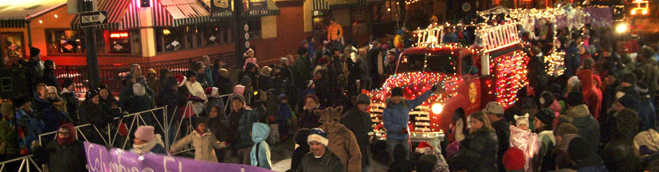 Desfile nocturno en Pearl Street: un camión de bomberos rojo cubierto de luces navideñas, multitudes de familias y una pancarta morada de Columbine al frente (recorte de banner de 942x247)